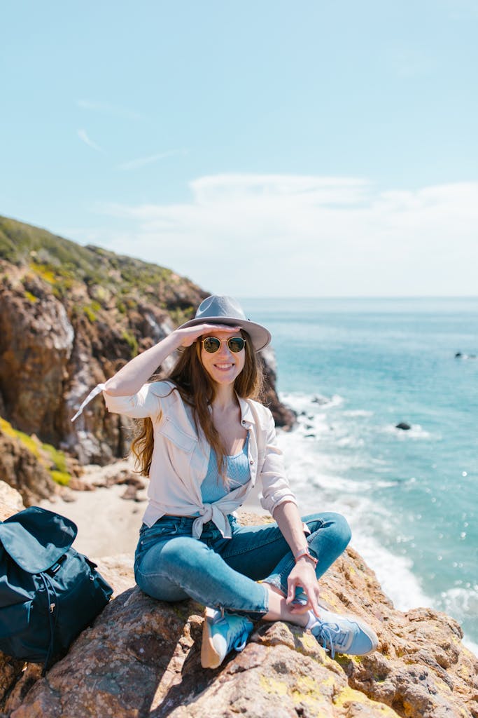 Young woman sitting on cliffs by the ocean, enjoying a sunny day outdoors.